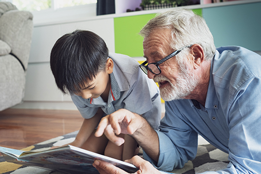 Older white man reading a book to a nine-year old Asian boy.